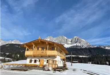 A cozy wooden house in a snowy landscape. Majestic mountains rise in the background under a clear blue sky.