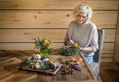 Eine Frau mit lockigem Haar sitzt an einem Tisch und arbeitet an Blumenarrangements. Neben ihr stehen zwei dekorative Gestecke aus bunten Blumen und Früchten.