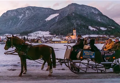 Eine Pferdeschlittenfahrt auf einer verschneiten Landschaft mit Bergen im Hintergrund. Die Passagiere genießen die winterliche Umgebung.