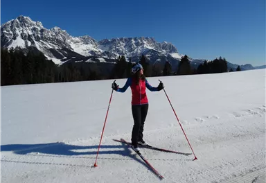 Eine Frau beim Skilanglauf in einer verschneiten Landschaft. Im Hintergrund sind majestätische Berge und ein klarer blauer Himmel zu sehen.
