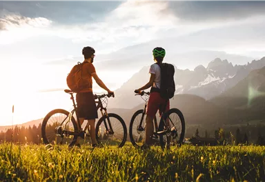 Zwei Radfahrer stehen auf einer Wiese und genießen den Sonnenuntergang über den Bergen. Die Aussicht ist malerisch und lädt zur Erkundung ein.