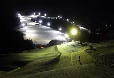 Eine beleuchtete Skipiste in der Nacht mit Schnee und einer klaren Sicht. Im Hintergrund sind Lichter, die die Strecke erhellen.