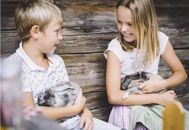 Two children are sitting next to each other and holding cute rabbits in their arms. They are laughing and having fun in a rustic setting.