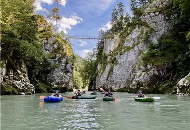 Eine Gruppe von Menschen paddelt auf einem ruhigen Fluss zwischen beeindruckenden Felsen. Im Hintergrund ist eine Brücke und der Himmel ist klar und blau.
