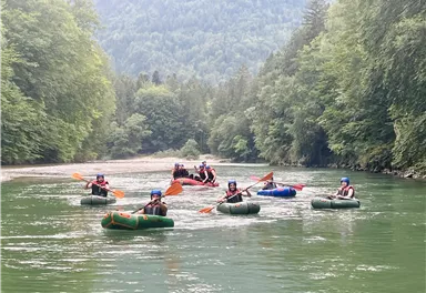 Eine Gruppe von Menschen paddelt in Gummibooten auf einem ruhigen Fluss. Umgeben von üppigem Grün und Bergen, herrscht eine entspannte Atmosphäre.