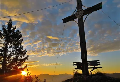 Ein großes Holzkreuz steht auf einem Hügel bei Sonnenuntergang. Im Hintergrund sind Berge und ein farbenfroher Himmel zu sehen.