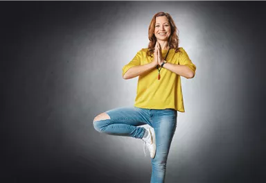 Eine lächelnde Frau steht auf einem Bein und hat die Hände in einer Gebetshaltung. Sie trägt ein gelbes Oberteil und graue Jeans.