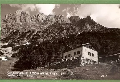 Eine Berglandschaft mit einer Berghütte im Vordergrund. Im Hintergrund erheben sich majestätische Gipfel unter einem bewölkten Himmel.
