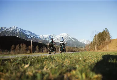 Zwei Personen fahren mit Fahrrädern auf einer Straße. Im Hintergrund sind schneebedeckte Berge und ein klarer Himmel zu sehen.