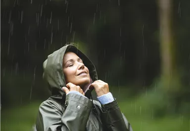 Eine Frau steht im Regen und genießt den Moment. Sie trägt eine grüne Regenjacke und schaut nach oben.