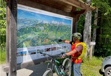 Ein Radfahrer steht vor einer Informationskarte in der Natur. Er zeigt auf die Karte, während sein Fahrrad neben ihm steht.