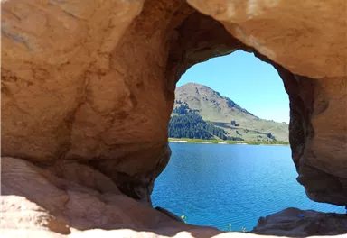 Ein Blick durch einen Felsen auf einen ruhigen See und sanfte Hügel im Hintergrund. Der Himmel ist klar und blau.