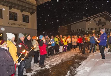 Eine Gruppe von Menschen steht in einer schneebedeckten Landschaft und hält Fackeln in der Hand. Es schneit und die Atmosphäre ist festlich und einladend.