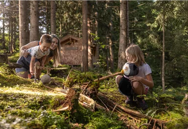 Kinder spielen im Wald und erkunden die Natur. Im Hintergrund ist ein Holzhaus zwischen den Bäumen zu sehen.