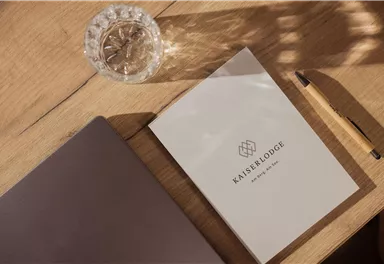 Ein elegantes Notizbuch mit dem Logo der Kaiserlodge liegt auf einem Holztisch. Daneben befinden sich ein Glas Wasser und ein Stift.