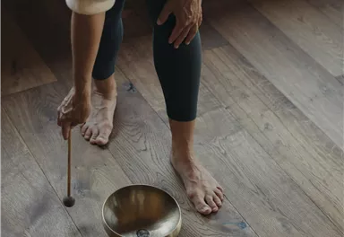 Eine Person steht barfuß auf dem Boden und spielt mit einem Schlägel auf zwei tibetischen Klangschalen. Die Umgebung wirkt beruhigend und harmonisch.