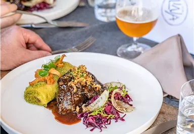 Ein Teller mit zartem Fleisch in einer dunklen Sauce, umgeben von buntem Gemüse. Im Hintergrund sind ein Glas Bier und weitere Teller zu sehen.