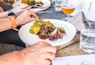 Ein Teller mit köstlichem Fleischgericht, garniert mit buntem Gemüse und frischen Kräutern. Im Hintergrund sind weitere Teller und Getränke sichtbar.