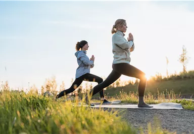 Zwei Frauen üben Yoga im Freien bei Sonnenuntergang. Die Umgebung ist grün und malerisch, was eine entspannende Atmosphäre schafft.