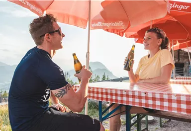 Ein Paar sitzt an einem Tisch mit rot-weißem Karomuster und genießt Getränke unter sonnigen Schirmen. Im Hintergrund sind Berge und eine schöne Aussicht sichtbar.