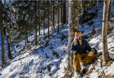 Eine Person sitzt entspannt in einem verschneiten Wald. Um sie herum stehen hohe Bäume und die Sonne scheint durch die Äste.