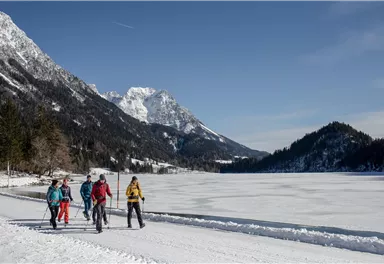 Eine Gruppe von Menschen wandert auf einem gefrorenen See neben schneebedeckten Bergen. Der Himmel ist klar und die Landschaft wirkt winterlich und einladend.