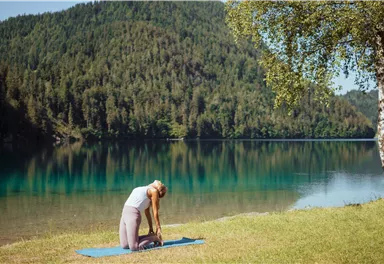 Eine Frau macht Yoga auf einer Matte am Ufer eines klaren Sees, umgeben von grünen Bäumen. Der Himmel ist blau und die Landschaft ruhig.
