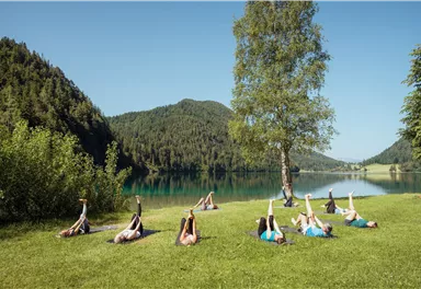 Eine Gruppe von Menschen macht Yoga auf einer Wiese am Ufer eines Sees. Im Hintergrund sind grüne Hügel und Bäume.
