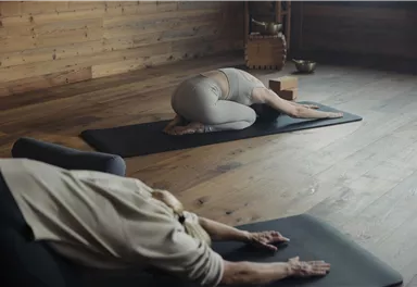 Eine ruhige Yogastunde in einem Holzraum. Zwei Personen praktizieren verschiedene Yoga-Positionen auf Matten.