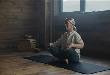 Eine Frau sitzt im Schneidersitz auf einer Yogamatte in einem gemütlichen Raum. Im Hintergrund sind Holzwände und einige Dekoartikel sichtbar.