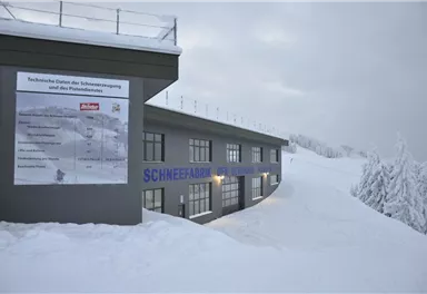 Ein Gebäude mit einer Schnee-Informationsstelle, umgeben von Schnee und winterlicher Landschaft. Der Himmel ist grau und bewölkt.