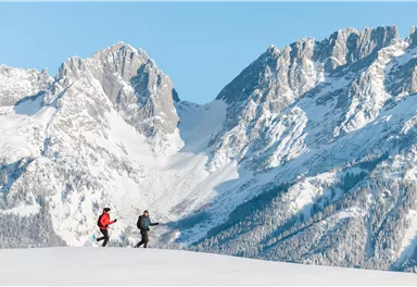 Zwei Personen laufen auf einer schneebedeckten Fläche in den Bergen. Im Hintergrund erheben sich beeindruckende, verschneite Berggipfel unter einem klaren, blauen Himmel.