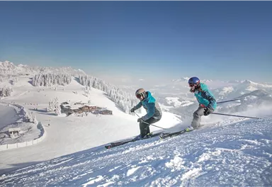 Zwei Skifahrer fahren die schneebedeckte Piste hinunter. Im Hintergrund sind verschneite Berge und eine Skihütte zu sehen.