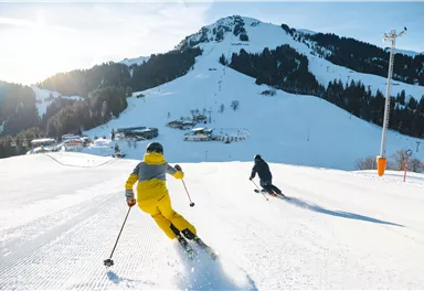 Zwei Skifahrer fahren die schneebedeckte Piste hinunter. Im Hintergrund sind Berge und ein klarer Himmel zu sehen.