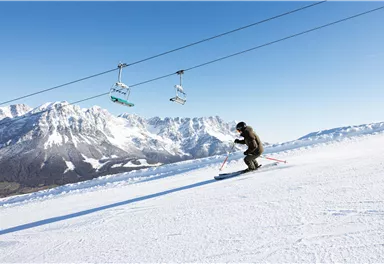 Ein Skifahrer fährt auf einer verschneiten Piste mit beeindruckenden Bergen im Hintergrund. Der Himmel ist klar und blau, und der Sessellift ist sichtbar.