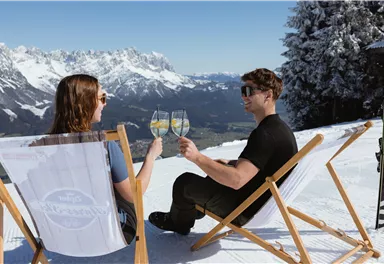 Ein Paar genießt Sekt auf einer Terrasse im Schnee. Im Hintergrund sind majestätische Berge und ein klarer blauer Himmel zu sehen.