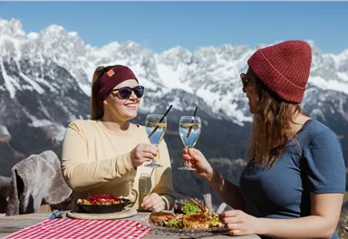 Zwei Frauen stoßen mit Gläsern auf einer Bergterrasse an. Im Hintergrund sind beeindruckende Berge und ein klarer Himmel zu sehen.