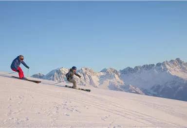 Zwei Skifahrer fahren auf einem schneebedeckten Hang. Im Hintergrund sind majestätische Berge und ein klarer blauer Himmel zu sehen.
