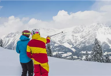 Zwei Skifahrer stehen im Schnee und schauen auf die Berge. Einer von ihnen zeigt mit einem Stock auf die Landschaft.