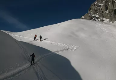 Drei Wanderer gehen über eine schneebedeckte Landschaft. Im Hintergrund sind Felsen und ein klarer blauer Himmel zu sehen.