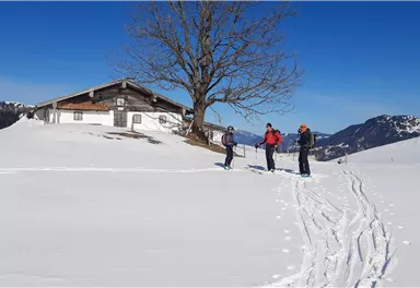 Eine verschneite Landschaft mit drei Personen, die im Schnee stehen. Im Hintergrund ist ein altes Haus und ein großer Baum zu sehen.