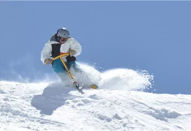Ein Skifahrer fährt mit Schwung die verschneite Piste hinunter. Der klare blaue Himmel bietet einen schönen Kontrast zum weißen Schnee.