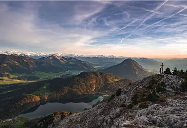 Eine beeindruckende Berglandschaft mit einem klaren Himmel und tiefgrünen Tälern. Im Hintergrund sind schneebedeckte Gipfel und ein glitzernder See zu sehen.