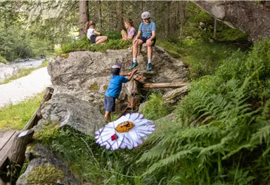 Eine Gruppe Kinder spielt in einem Waldgebiet. Im Vordergrund ist eine große, bunte Blume zu sehen.
