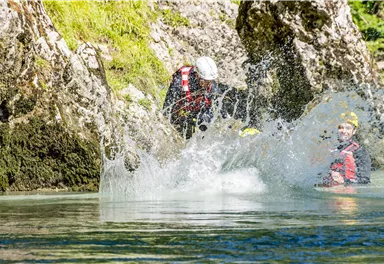 Eine Person spritzt Wasser, während sie im klaren Wasser steht. Im Hintergrund sind Felsen und eine weitere Person zu sehen.