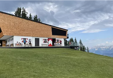 Ein modern gestaltetes Gebäude mit Holzverkleidung, umgeben von grünem Rasen. Im Hintergrund sind majestätische Berge und ein bewölkter Himmel zu sehen.