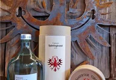 Eine elegante Flasche Edelbrand steht neben einer runden Holzverpackung und einer dekorativen Dose. Im Hintergrund ist ein rustikales Metall-Symbol zu sehen.