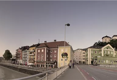 Eine malerische Straßenszene mit bunten Häusern entlang eines Flusses. Im Hintergrund ist ein Hügel mit einer alten Festung zu sehen.