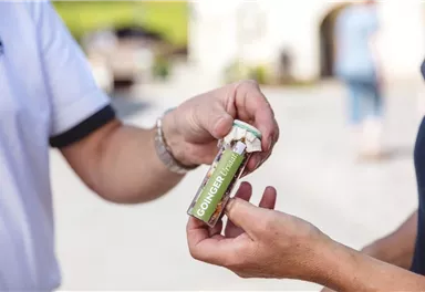 Eine Person reicht einer anderen ein kleines Röhrchen mit der Aufschrift "GONDER". Das Röhrchen ist in einer natürlichen Umgebung zu sehen.
