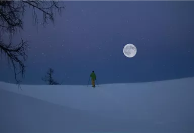 Eine Person in grünem Outfit steht im verschneiten Gelände unter einem klaren Nachthimmel. Der Vollmond leuchtet hell über der Winterlandschaft.